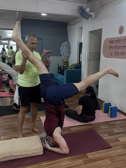 An instructor provides support to a student practicing a variation of Sirsasana (Headstand), ensuring stability and safety while exploring this advanced inversion.