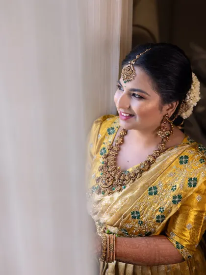 A radiant bride-to-be, her smile lit by the soft light from a window. This candid portrait captures her happiness and the beautiful details of her golden jewelry.