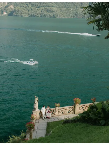 A wide, scenic shot of a couple on a beautiful stone terrace overlooking the vibrant blue waters of Lake Como, Italy. The image captures the grandeur and serenity of the location.