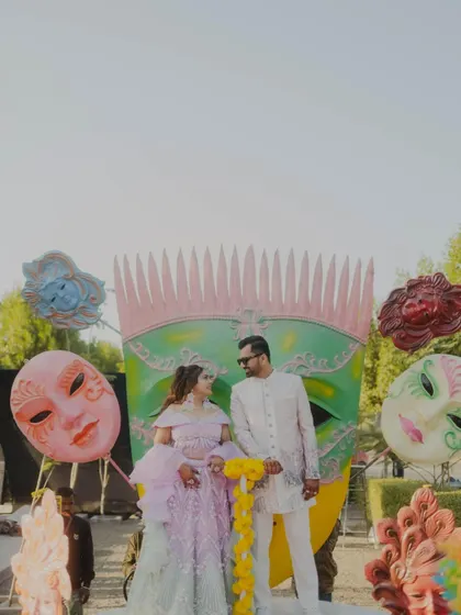 A quirky and fun shot of the couple posing with large carnival masks. This image perfectly captures the playful and festive theme of their pre-wedding celebration.