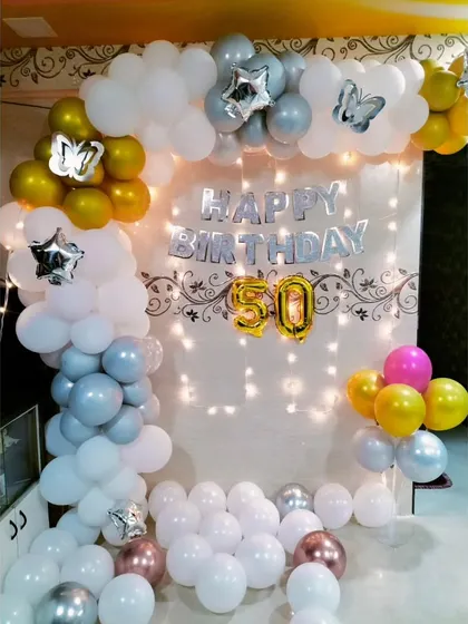 A beautiful home setup for a 50th birthday, using a white, gold, and silver balloon arch with fairy lights and butterfly accents.