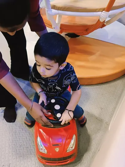 A toddler enjoys a ride in a small toy car with a parent's help, a fun gross motor activity.