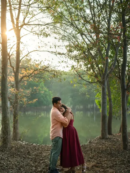 A couple embraces by a lake at sunset, surrounded by trees. This romantic outdoor portrait uses the beautiful evening light to create a warm, intimate, and picturesque scene.