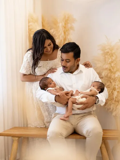A lovely family portrait with the father seated and holding both twins. The mother stands behind him with her hands on his shoulders, looking down at her new babies with a loving expression.