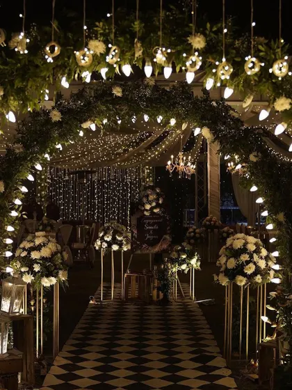 A glamorous Sangeet passage with an arch of greenery and white flowers, lit by hanging bulbs. The checkered floor and fairy light ceiling create a chic, party-ready atmosphere.