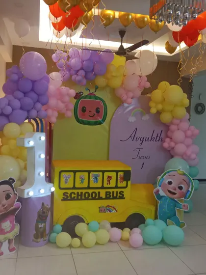 A home-based Cocomelon first birthday party. The setup includes a yellow school bus prop, character cutouts, and a pastel balloon arrangement.