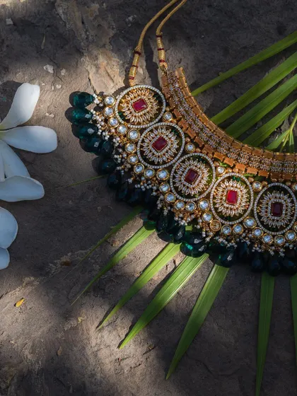 A stunning detail shot of a traditional bridal necklace, laid out on a palm leaf. The intricate design with rubies, emeralds, and pearls showcases the exquisite craftsmanship of Indian wedding jewelry.