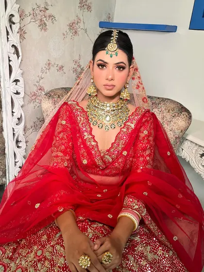 A regal portrait of a bride in a classic red lehenga. The makeup features sharp, defined eyes and a perfectly sculpted base, paired with a heavy kundan necklace to create a look of pure bridal opulence.