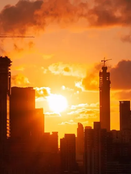 The sun sets between the silhouettes of Mumbai's buildings, with cranes visible on under-construction towers, all under a fiery orange sky.