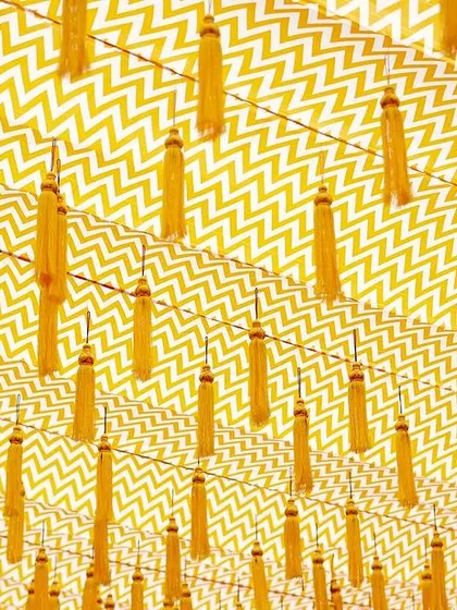A creative ceiling treatment using a chevron-patterned fabric with hanging yellow tassels, adding a modern graphic element to the traditional Haldi decor.