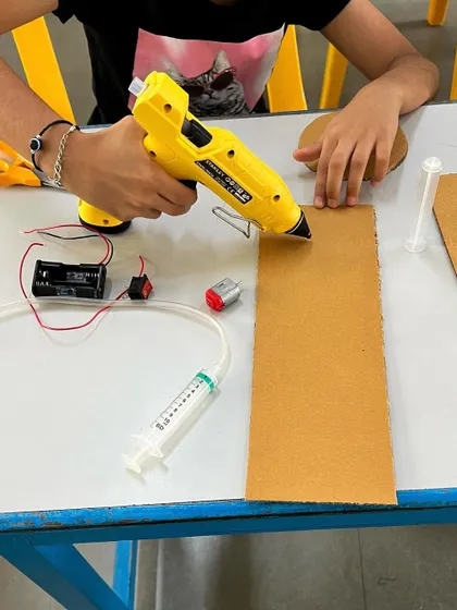 A student uses a glue gun to assemble the cardboard frame for a hydraulic disc brake model. This project requires careful construction to ensure the mechanical parts work correctly.