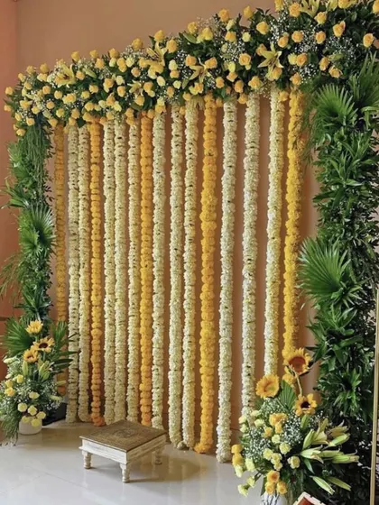 A simple and classic Haldi photo backdrop made of yellow and white flower strings, framed with green leaves. This is a timeless choice for home functions.