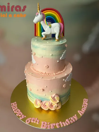A whimsical two-tier unicorn cake featuring pastel rainbow frosting and a beautifully sculpted unicorn under a colorful rainbow. The delicate clouds and piped swirls add to the magical, dreamy feel of this sixth birthday cake.