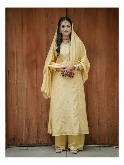 A full-length shot of the bride in a simple yet elegant yellow salwar suit, standing against a rich wooden background.