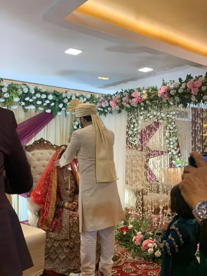 A candid moment from a Nikah ceremony. The groom is seen with the bride, who is behind a veil, against the backdrop of a beautiful floral partition.