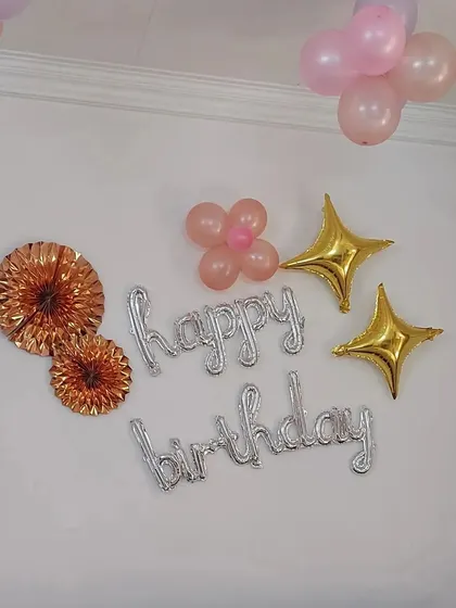 A simple and sweet wall decoration for a birthday. It includes silver "Happy Birthday" foil letters, star-shaped foil balloons, and small clusters of pink balloons.
