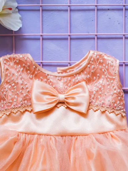 A lovely peach-colored party dress featuring an intricately embroidered bodice and a sweet satin bow. The combination of textures adds depth and elegance to the design.