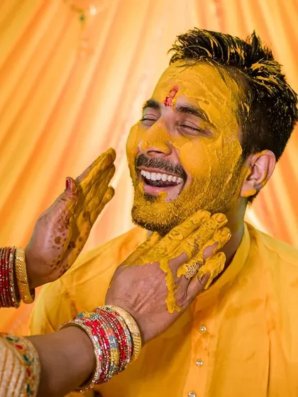 A close-up of the groom smiling as turmeric is gently applied to his face. This shot captures a softer, more traditional moment amidst the fun.