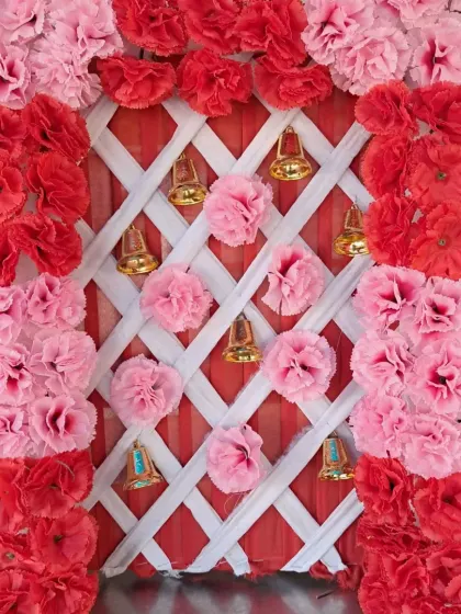 A compact backdrop design featuring a frame of red and pink artificial carnations. The background uses a white lattice pattern over red fabric, decorated with hanging golden bells and pom-poms.