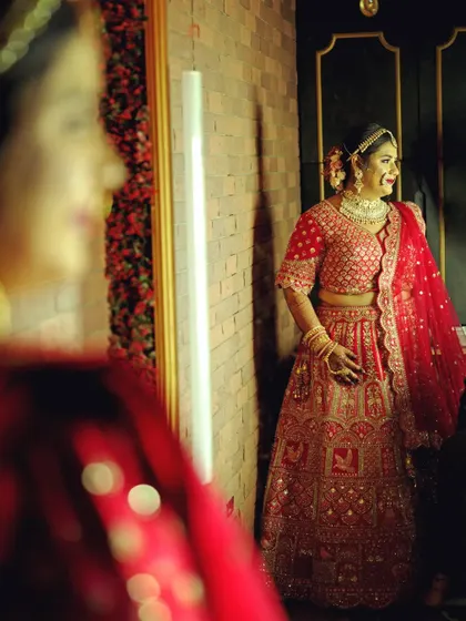 A candid shot of the bride admiring her reflection. This photo captures a moment of happy anticipation, with her radiant smile and elegant red lehenga filling the frame.