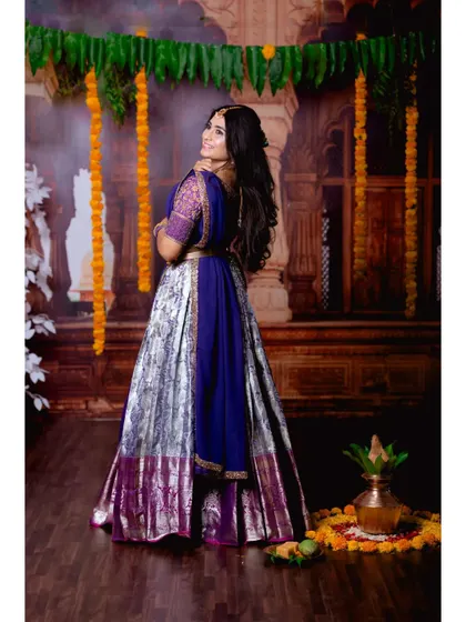 A back view of the purple half-saree, showcasing the full lehenga skirt and the traditional backdrop decorated with marigolds.