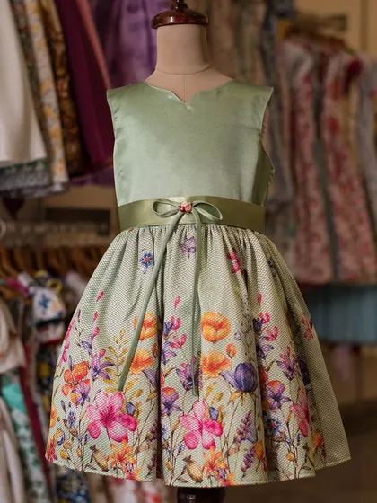 A sophisticated dress in sage green, featuring a solid satin bodice and a beautiful floral border print on the skirt. The thin belt with a small bow adds a delicate finishing touch.