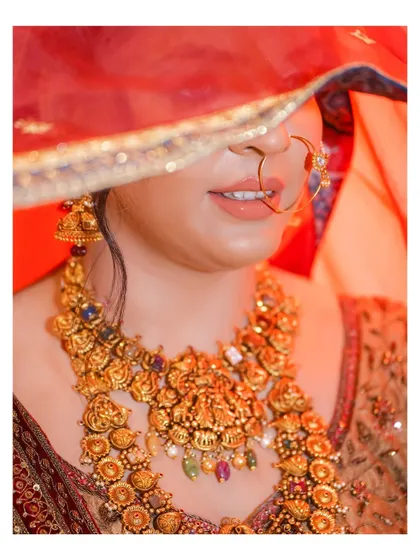 An artistic close-up of the bride, her face partially hidden by her red veil. The focus is on her magnificent temple jewelry, showcasing its intricate design.