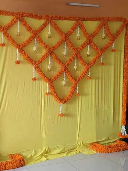 A beautiful and simple backdrop made entirely of fresh marigold flowers. The geometric lattice pattern with hanging bells is a traditional and fragrant choice for an at home Haldi or Pooja ceremony.