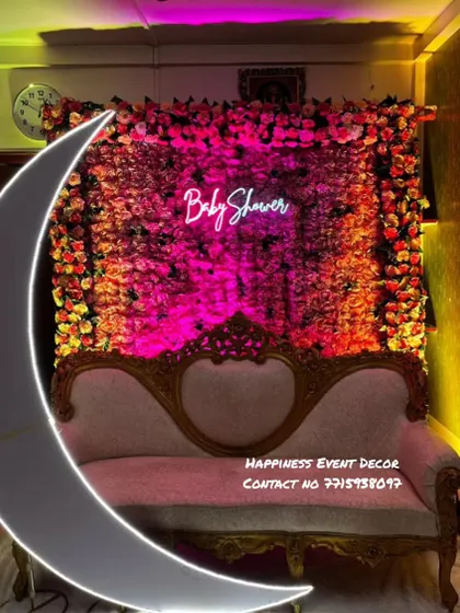 A close-up of a baby shower backdrop featuring a dense wall of pink and orange flowers. The 'Baby Shower' neon sign and a classic sofa create a perfect photo corner for the mom-to-be.