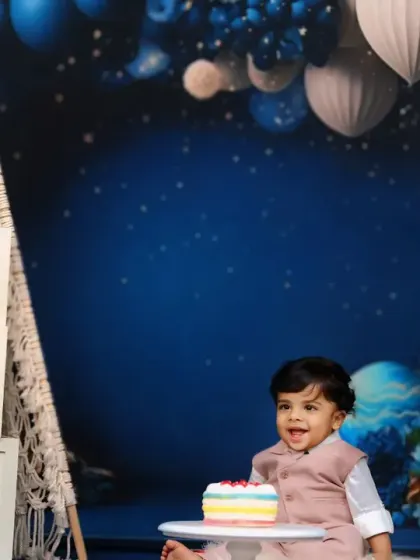 A little boy celebrates his first birthday with a rainbow cake, set against a celestial blue backdrop with a teepee and 'ONE' blocks.