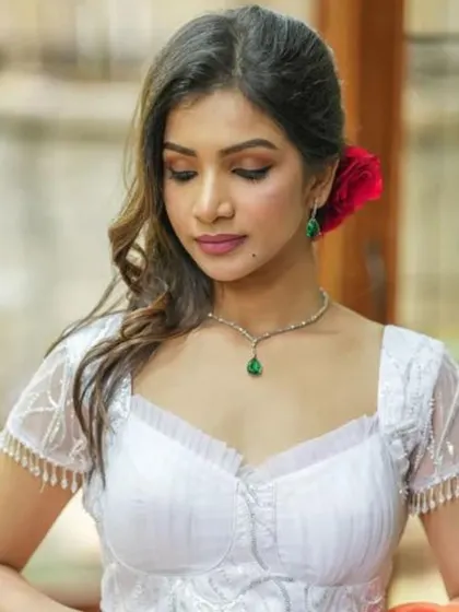 A model wears a delicate white blouse with fringe details on the sleeves, accessorized with a simple emerald pendant necklace.