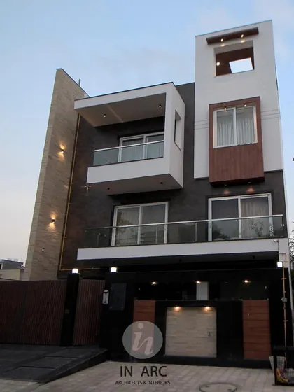A full view of a modern residence elevation. The design plays with geometric forms, contrasting white planes with textured stone walls and warm wood accents to create a balanced and dynamic facade.