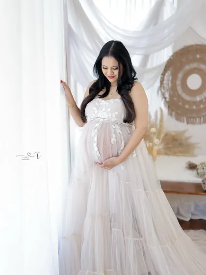 A beautiful and serene portrait. The mom-to-be looks down at her bump with a gentle smile, dressed in a delicate white lace gown in a boho-chic setting.