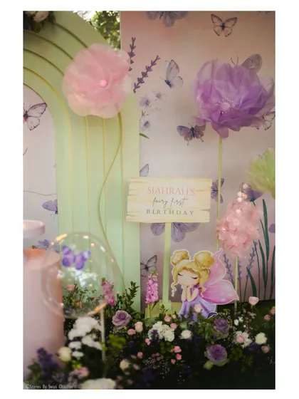 A close-up of the fairy garden decor, featuring a personalized sign, a cute fairy cutout, and large, delicate paper flowers against a butterfly-print backdrop.