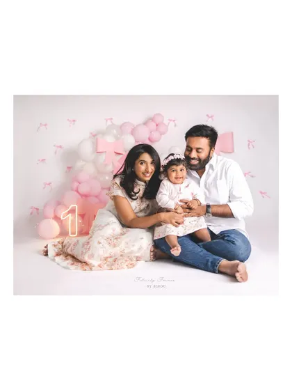 A smiling family celebrating their daughter's first birthday. The pink and white balloon decor, along with a glowing "1" sign, creates a perfect party atmosphere in the studio.