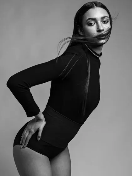A high-fashion, editorial-style bodysuit shot in black and white. The dynamic pose and wind-blown hair create a sense of power and movement, while the lighting sculpts the body beautifully.