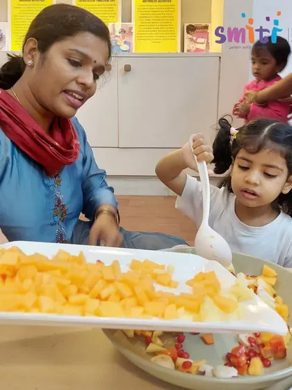 A teacher helps a young girl add chopped fruit to a large bowl to make a fruit salad. This activity teaches practical skills and the joy of preparing food for others.
