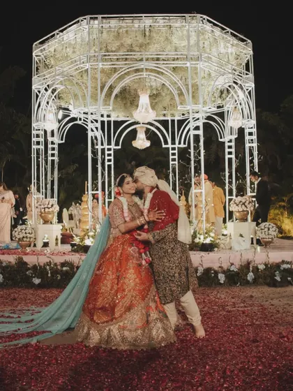 The couple shares a kiss in front of their beautiful wedding mandap, a perfect moment framed by the elegant structure and a carpet of scattered rose petals.