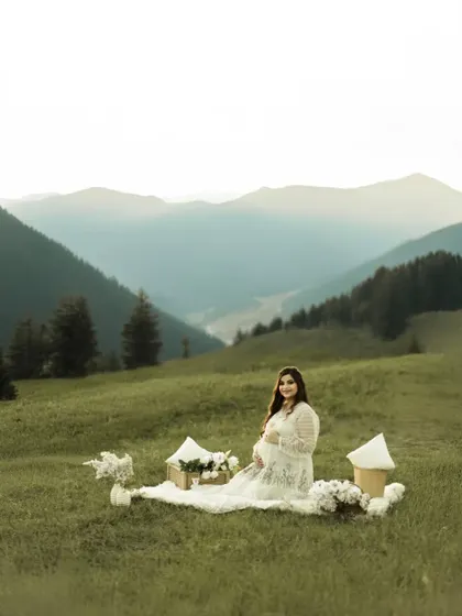 A breathtaking portrait in the mountains. The mother-to-be sits on a picnic blanket in a vast green meadow, with layers of mountains stretching out behind her, creating a sense of peace and grandeur.