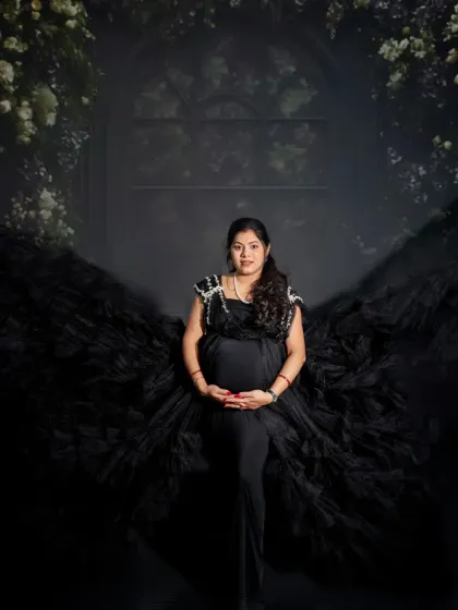 A powerful and angelic solo portrait. The mom-to-be stands in a black gown with a wing-like train, framed by a dark floral arch, creating a striking image.
