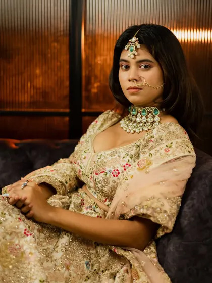 A full-length bridal portrait in an elegant indoor setting. The bride is seated, showcasing her magnificent embroidered lehenga and stunning Polki jewelry.