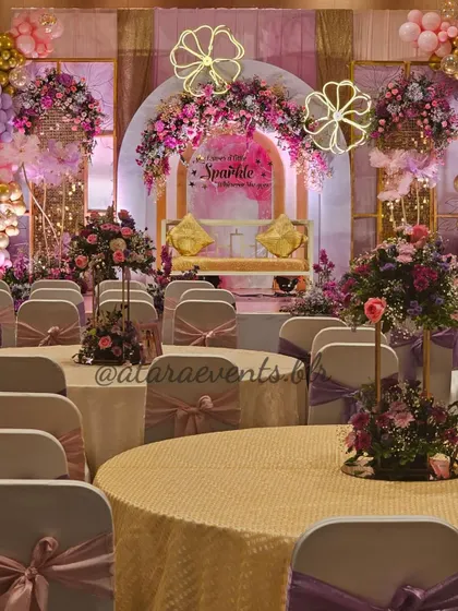 The elegant seating arrangement εταιρεία an Aqeeqah ceremony. The tables are beautifully decorated, and the chairs have lovely sashes, all complementing the grand stage setup in the background.