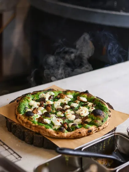 A steaming hot Fungi pizza on the prep counter. The rising steam is a testament to its freshness, straight from the intense heat of the wood oven.