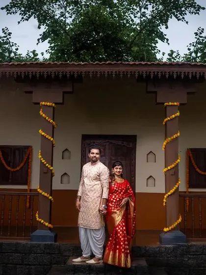 A classic portrait of a couple standing on the steps of a traditional Maharashtrian house, decorated with marigold flowers.