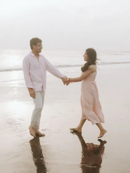 Ritu and Tarush holding hands as they walk along the shoreline. The soft sunset light and their reflections in the wet sand create a dreamy, romantic atmosphere for this pre-wedding photograph.