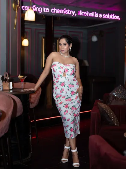 A full-length fashion portrait taken in a chic cafe. The neon sign and plush seating add to the trendy, urban vibe of this influencer-style shoot.