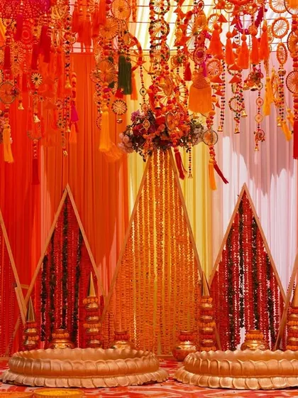 A closer look at the Haldi ceremony stage. The setup features traditional brass 'urli' bowls for the rituals, set against a backdrop of shimmering gold and orange fabrics and intricate hangings.