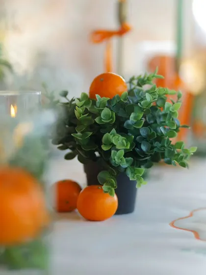 A small potted plant with a fresh orange placed on top serves as a simple, fresh, and on-theme decorative element for the tables.
