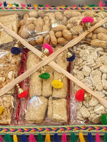 A square tray hamper for Lohri, filled with an assortment of traditional winter sweets like gajak and chikki, decorated with colorful pom-poms.