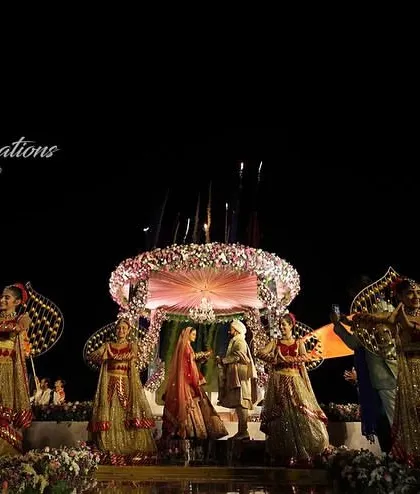 A wide-angle shot of a beach wedding Jaimala, capturing the entire magnificent setup. The stage, performers, and fireworks come together to create a scene of epic proportions.